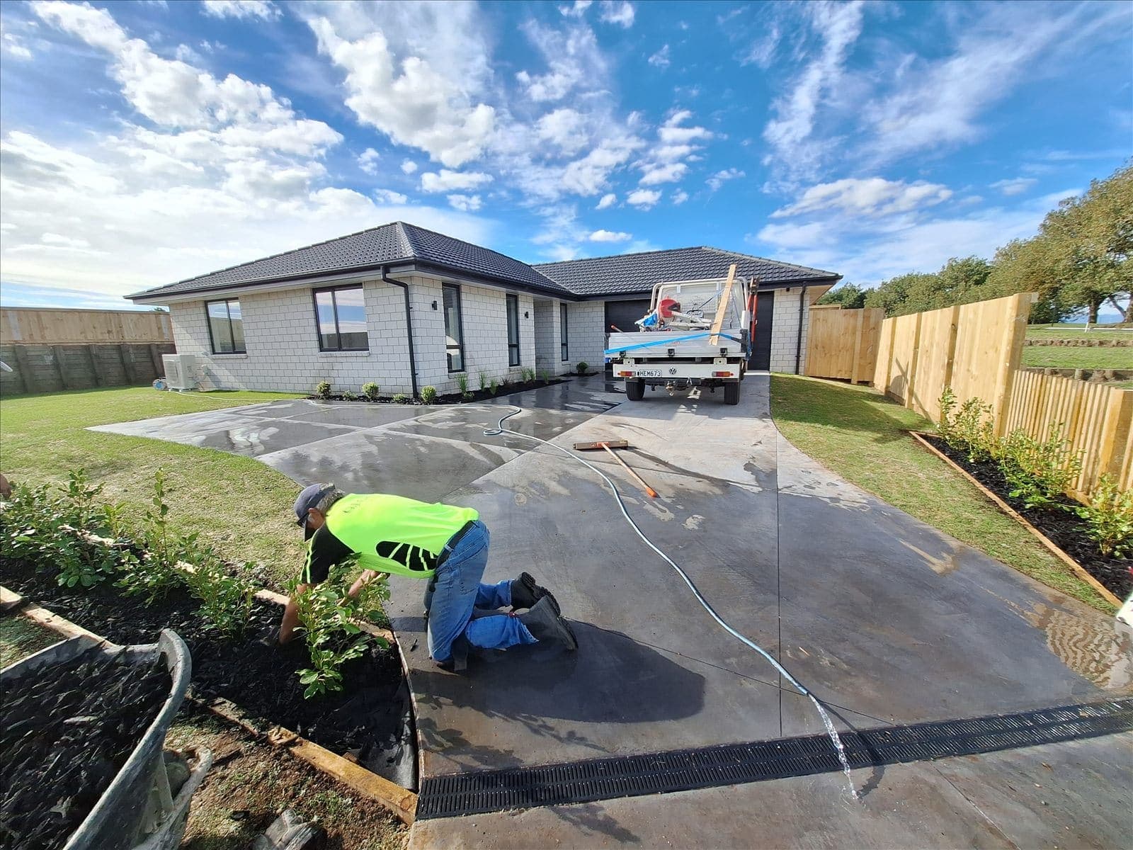 A freshly poured concrete driveway at an urban new-build home in Hamilton, a Tensen Landscapes crew member finishing the garden edges beside it